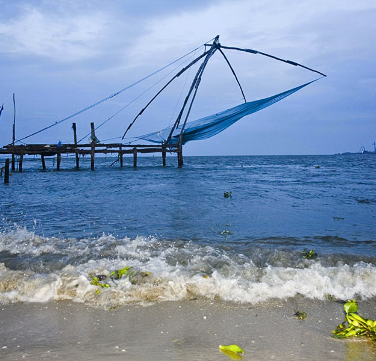 A large, traditional kerala's fishing net is suspended over the water, attached to a wooden structure. The ocean waves gently lap at the sandy shore as houseboats drift by, offering scenic views. A few green leaves are scattered nearby. The sky is overcast with shades of blue and gray, indicating an approaching evening.