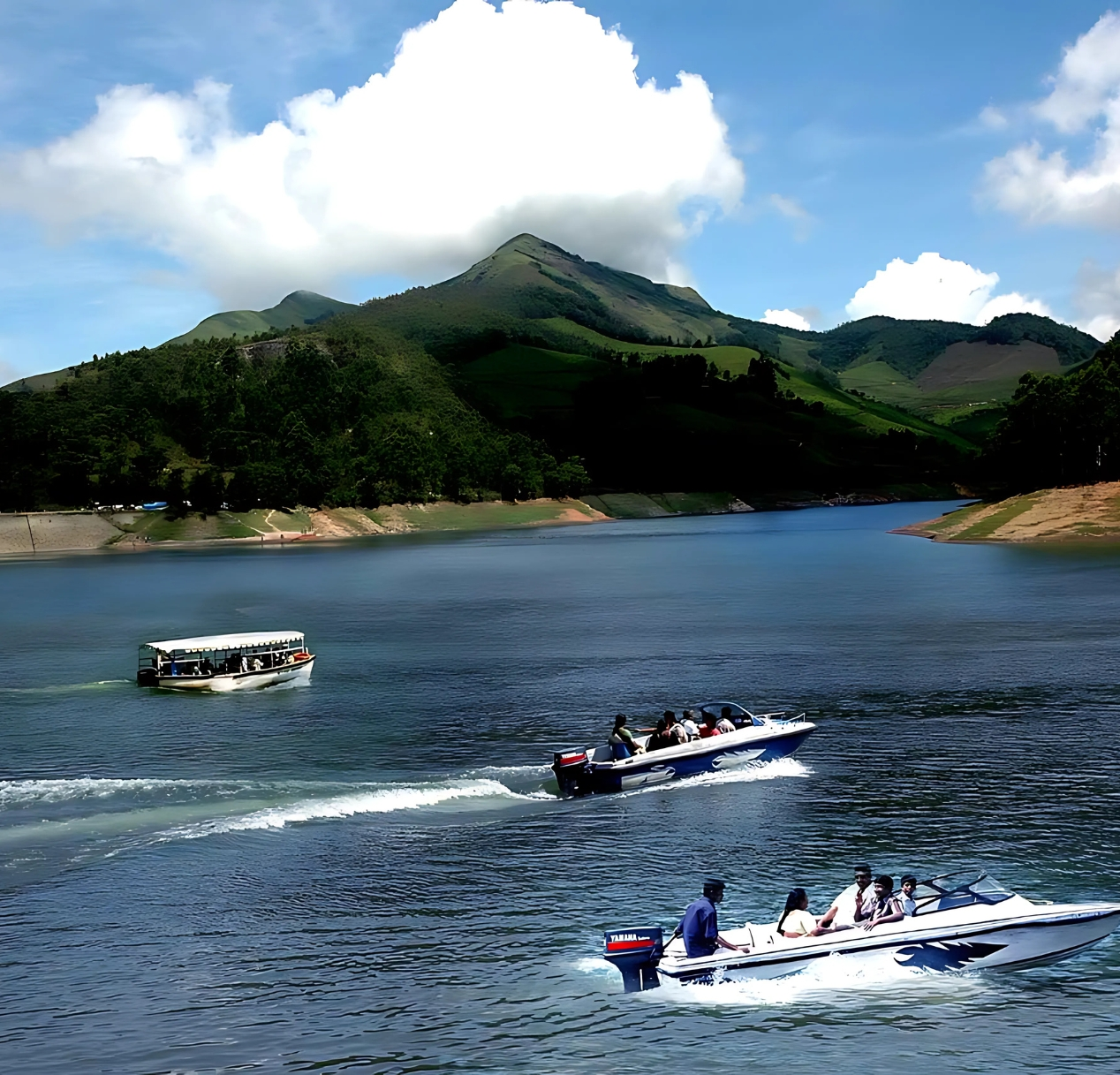 A tranquil lake surrounded by lush green hills under a clear blue sky. Three motorboats glide on the water, with one carrying passengers. The scene is peaceful, highlighting the natural beauty akin to 4 day Kerala tour packages, with reflections visible on the waters surface.