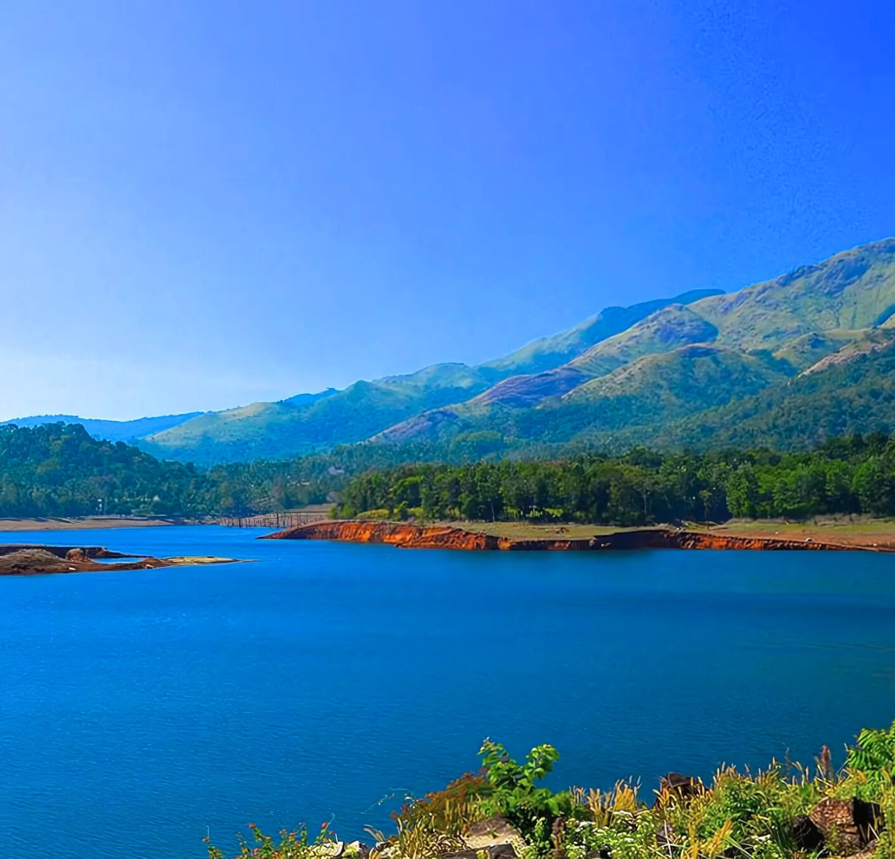 A serene lake with deep blue water reflects the clear sky, reminiscent of the scenic views often seen on a 4 day Kerala tour package. Surrounding the lake are lush, green hills and mountains that stretch into the distance. Light green vegetation graces the foreground, as bright sunlight bathes this peaceful and natural scene.