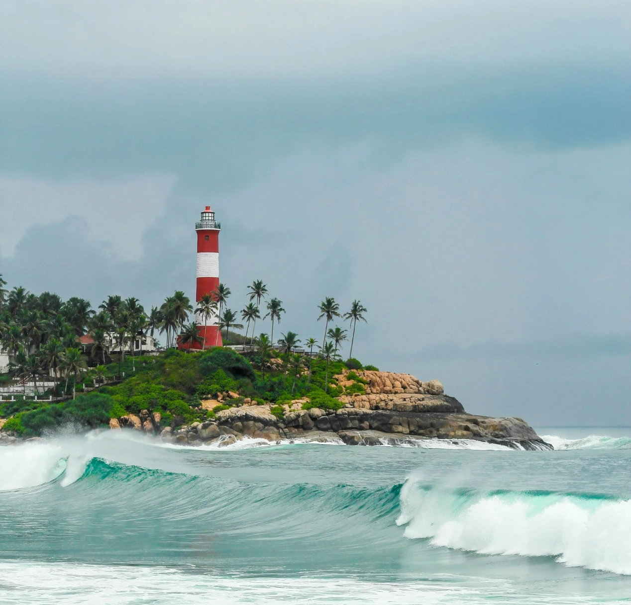 A towering red and white striped lighthouse stands on a rocky, palm-covered hill beside the ocean, offering scenic views. Large waves crash against the rocks as an overcast sky with dark clouds hints at an incoming storm, creating a dramatic coastal scene perfect for any 4 day Kerala tour package.