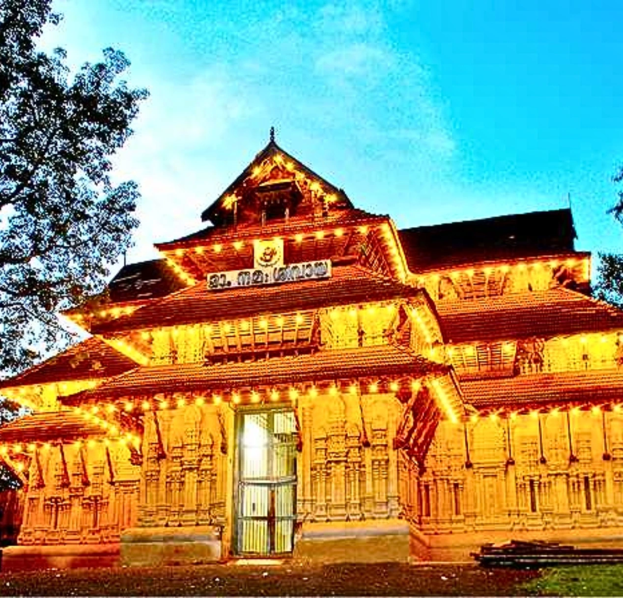 A beautifully lit traditional Thrissur temple stands against a dusky blue sky, reminiscent of the scenic views offered in a 4 day Kerala tour package. The building features intricate wood carvings and tiered roofing adorned with warm golden lights. Trees frame the scene, adding to the tranquil evening ambience.