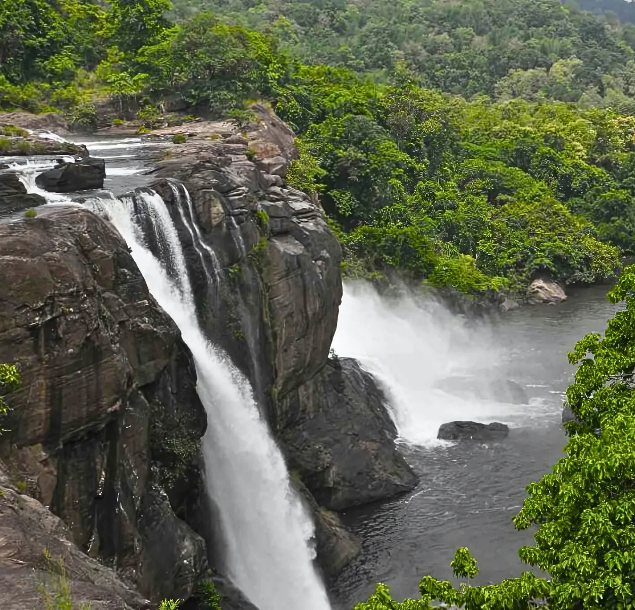 A majestic waterfall cascades down a rocky cliff surrounded by lush green foliage, offering scenic views reminiscent of a 4 day Kerala tour package. The water crashes into a serene pool below, creating misty spray. Dense forest stretches out into the background, emphasizing the waterfalls height and natural beauty.