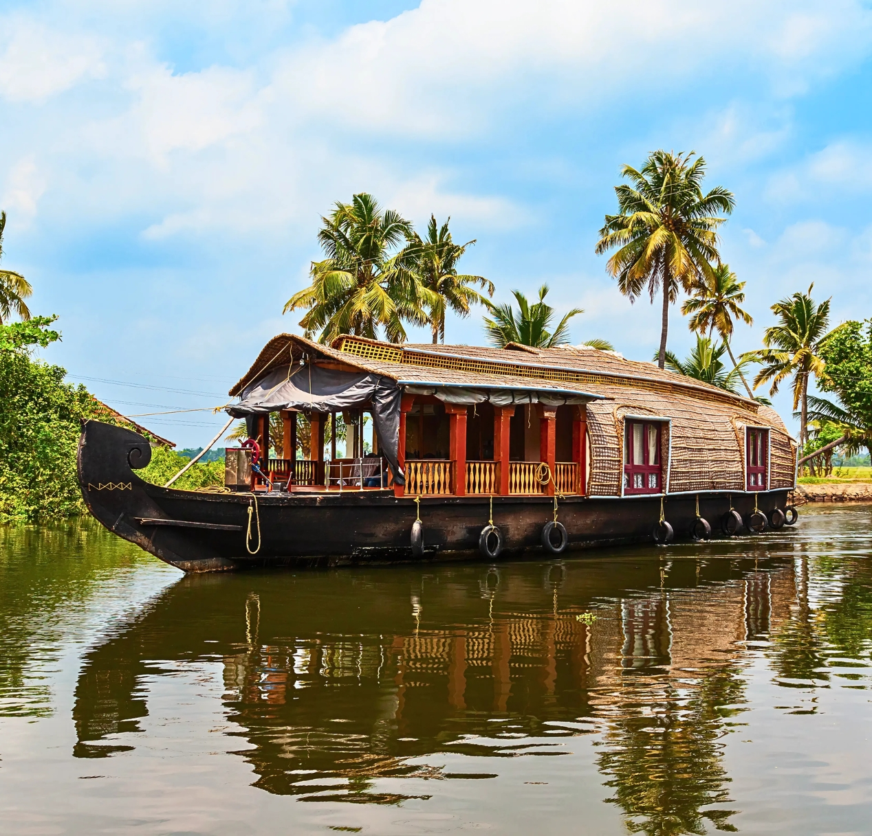 A traditional wooden houseboat with a thatched roof floats on a tranquil waterway, offering scenic views of lush greenery. Palm trees line the shore beneath a partly cloudy sky, reflecting in the water. Ideal for a 4 day Kerala tour package, the boat has open windows and an inviting appearance.