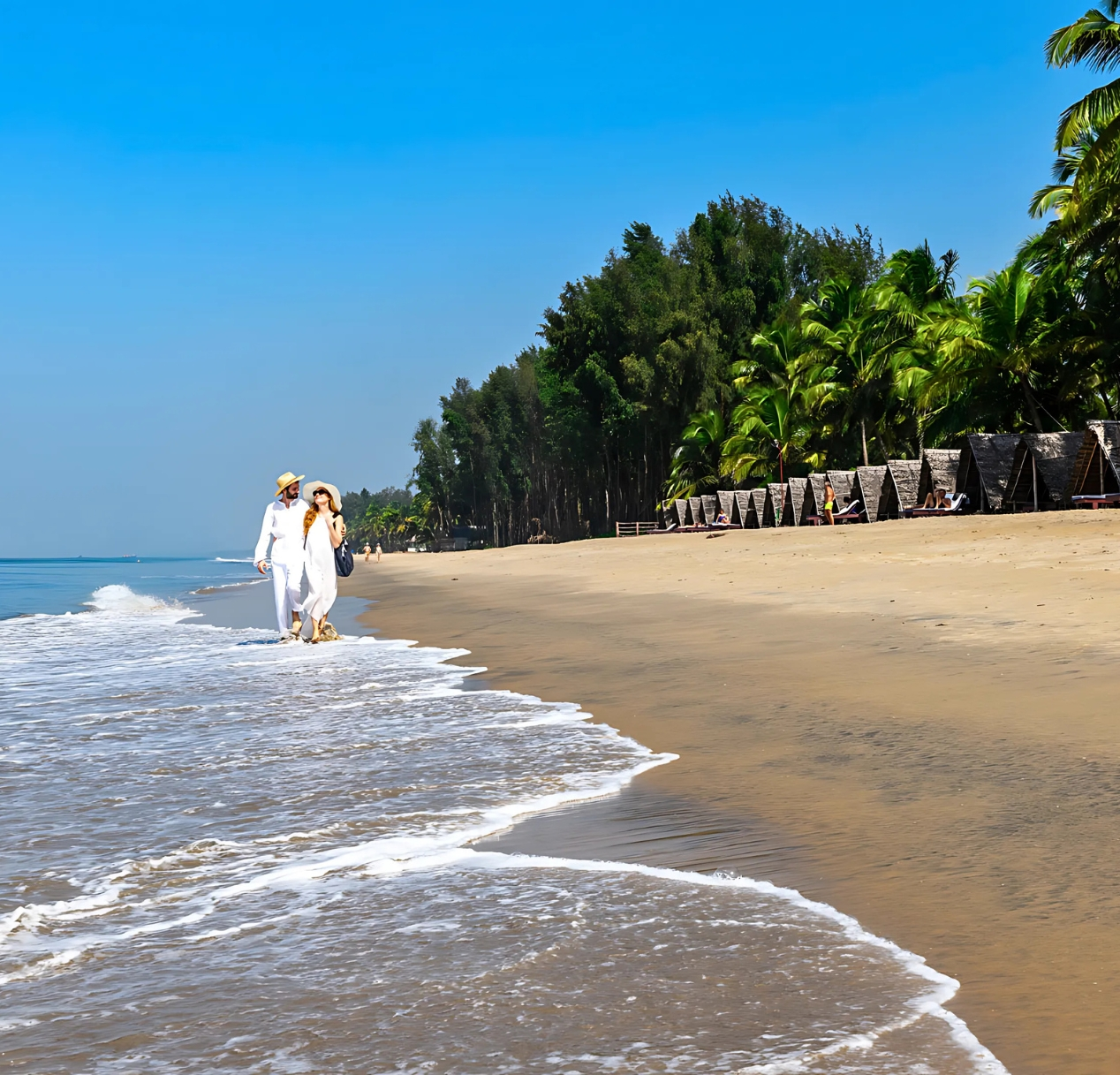 A couple dressed in white, wearing sun hats, walks hand in hand along a serene beach. Gentle waves lap at their feet. The idyllic 4 day Kerala tour package includes lush palm trees and beach huts. The clear blue sky enhances the tranquil and picturesque scene.