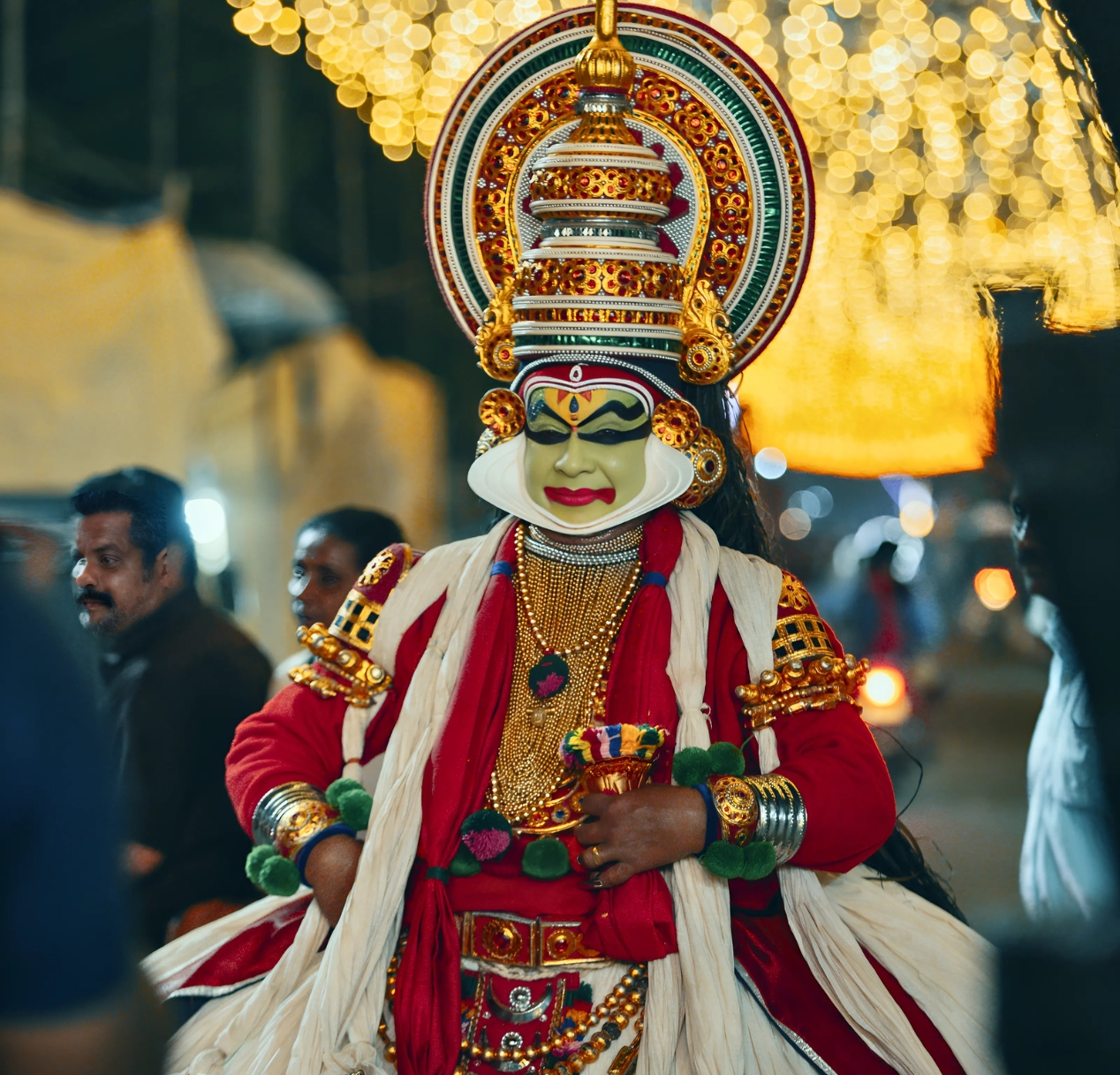 A person dressed in traditional Kathakali attire, featuring a vibrant red and white costume, ornate headdress, and elaborate face paint, stands in a festive, illuminated street setting. The scenic views are enhanced by glowing lights, creating a lively atmosphere that echoes the charm of a 4 days Kerala tour package.