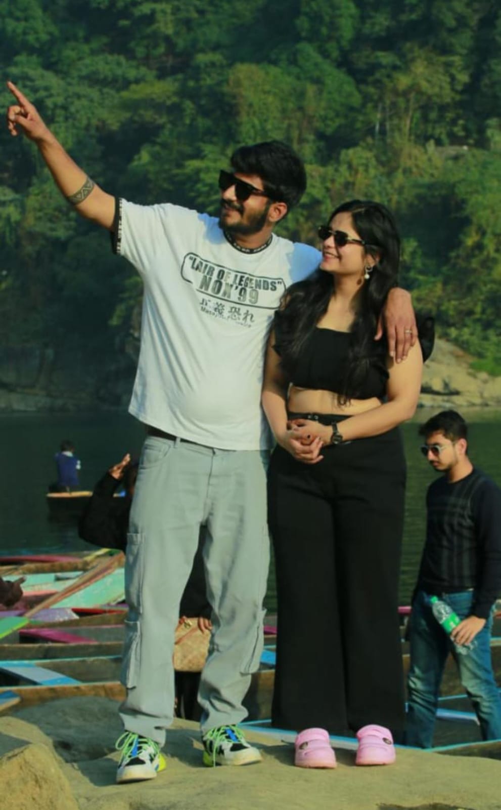 A couple wearing sunglasses poses happily by a lake, surrounded by greenery and colorful boats Dawki River meghalaya. The man wears a white T-shirt and gray pants, and the woman is in a black outfit. Other people are visible in the background.