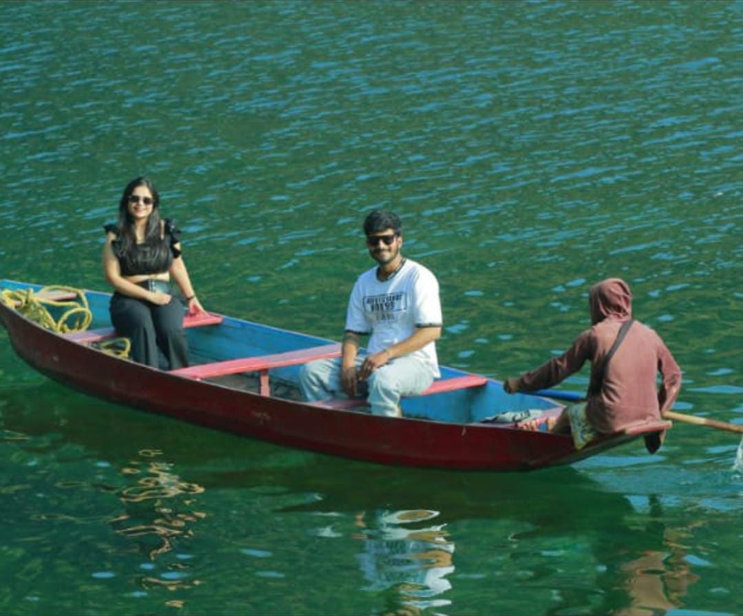 Three people in a colorful canoe on a calm green Dawki River meghalaya: a woman and a man seated facing forward, and a person in a hood operating the boat. They are enjoying a peaceful day on the water.