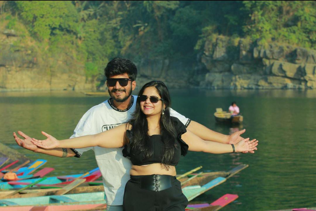 A man and woman happily pose for a photo, standing with arms outstretched beside a scenic Dawki River meghalaya . Colorful boats float in the water, and a rocky, tree-lined shore is visible in the background under a clear sky.