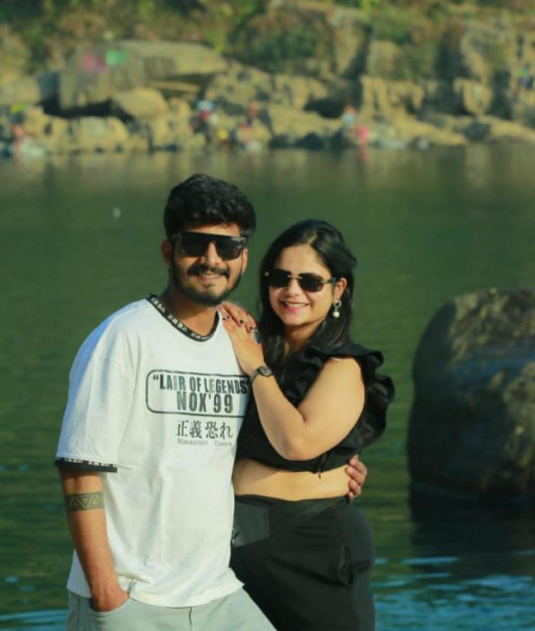 A couple poses happily in front of a tranquil body of water in meghalaya, surrounded by rocks and distant people. The man wears sunglasses and a white t-shirt, while the woman is in a black outfit and sunglasses. They stand close together, smiling.