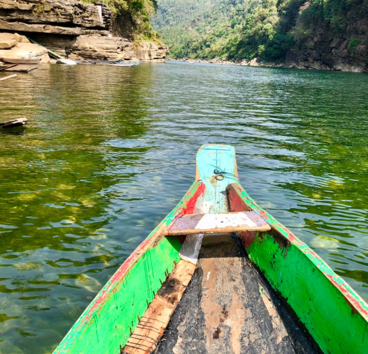 A colorful canoe with a green and red bow glides on a clear Dawki River surrounded by lush in meghalaya, rocky hillsides. The water reflects the vibrant greenery of the landscape, creating a serene and natural setting.