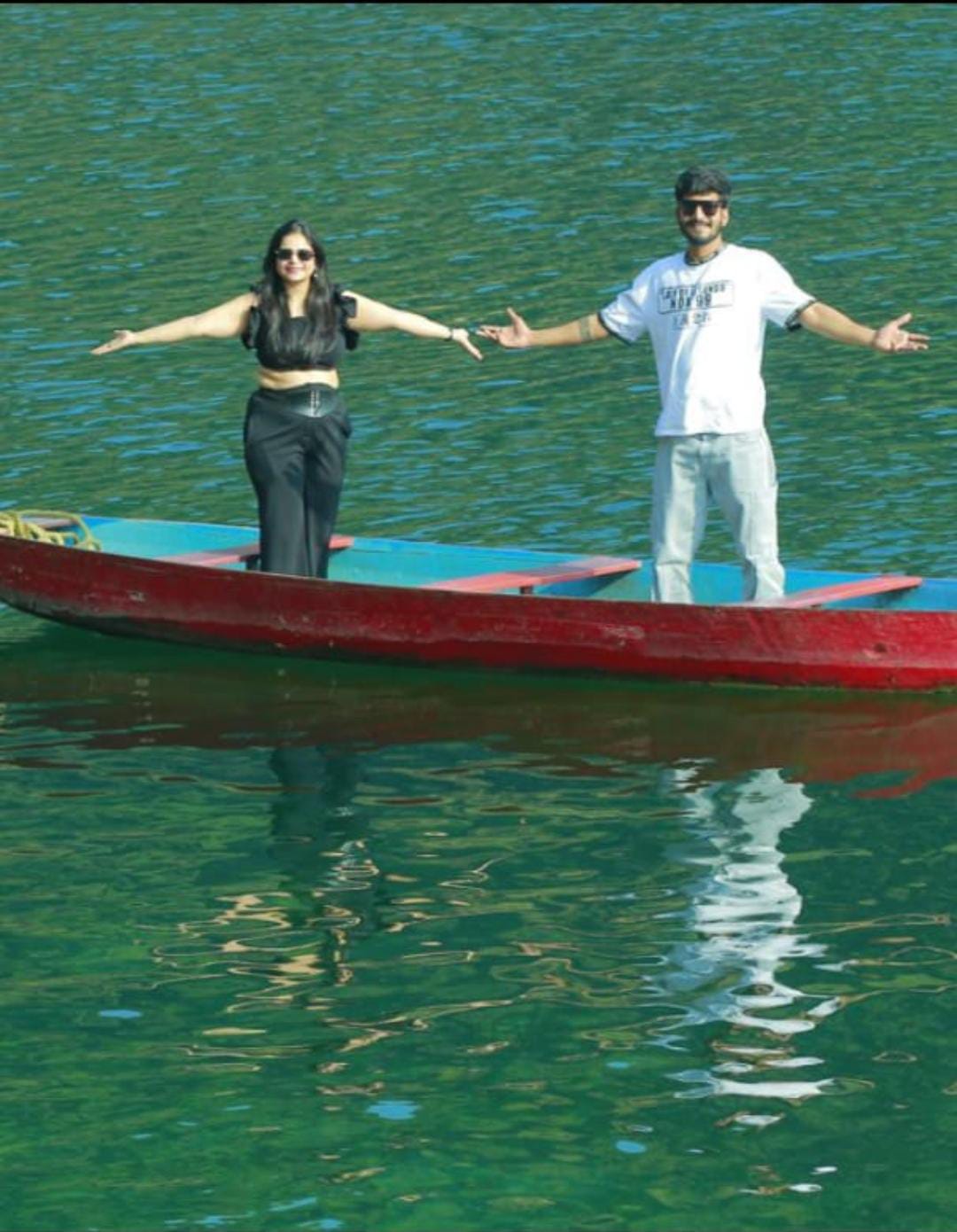 A woman and a man stand with arms outstretched on a narrow red boat, floating on a calm green lake meghalaya . Both are smiling and wearing casual outfits, with the woman in black and the man in white and blue.