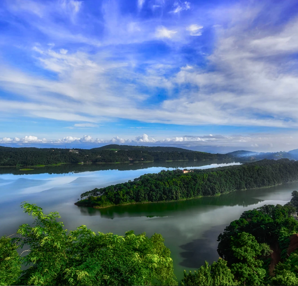 A scenic view of a meghalaya's lush green landscape with Umiam Lake surrounded by dense forests. The sky is bright blue with scattered clouds, casting reflections on the calm water. Hills are visible in the distance.