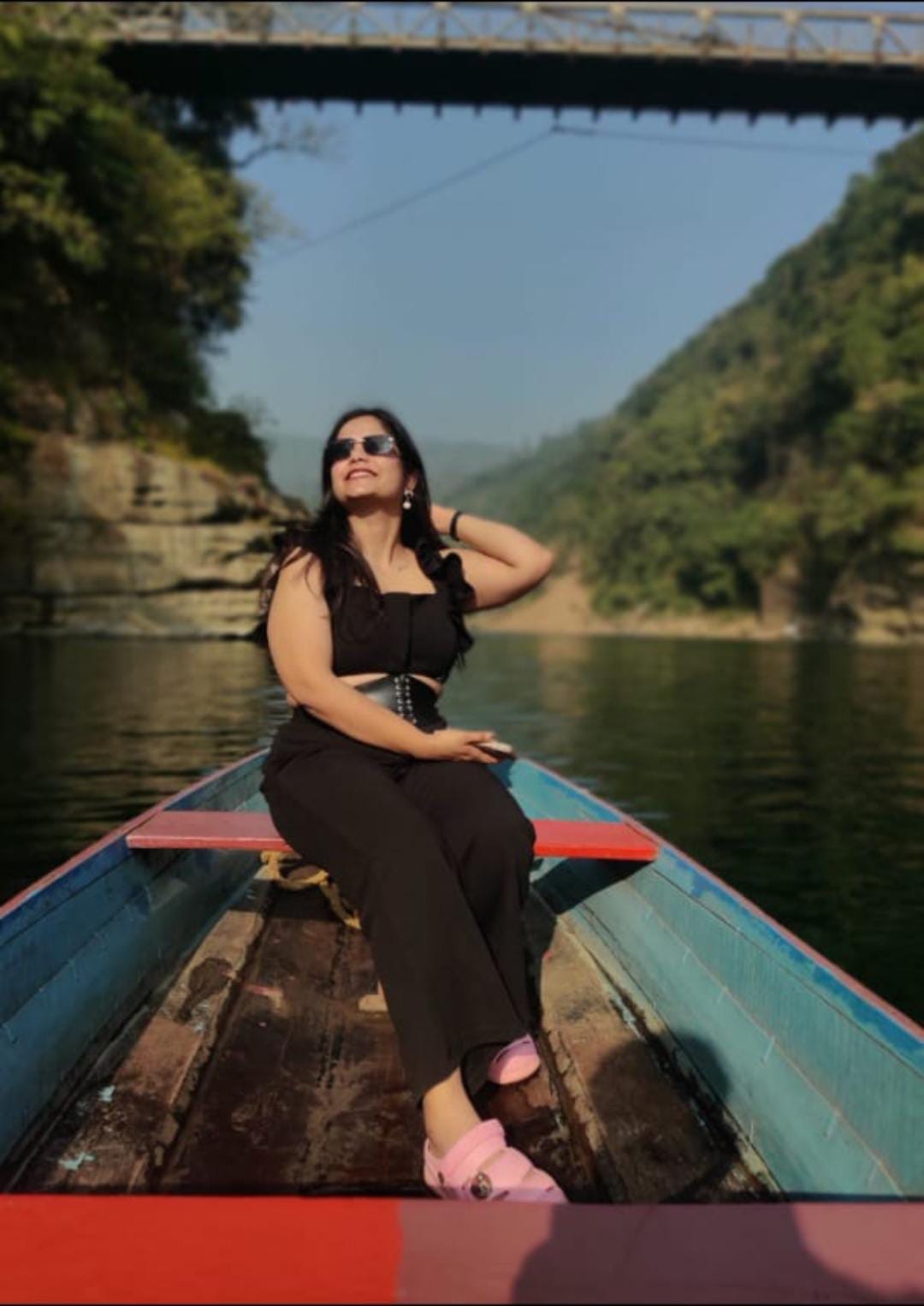 A woman in sunglasses and a black outfit sits on a boat in a calm Dawki River meghalaya, surrounded by lush green hills under a clear blue sky.