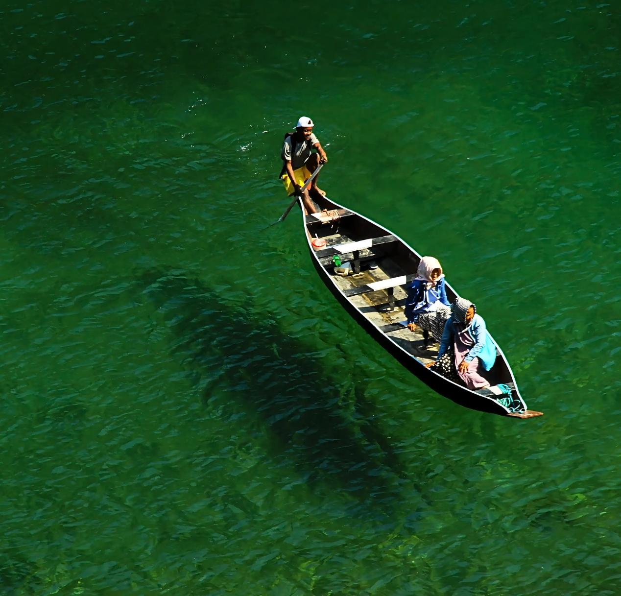 A small wooden boat with three people is gliding over clear, emerald-green water in meghalaya Dawki River. The boats shadow is visible on the riverbed. The people are wearing colorful clothing and hats, and one is rowing. The scene captures tranquility and the vividness of the water.